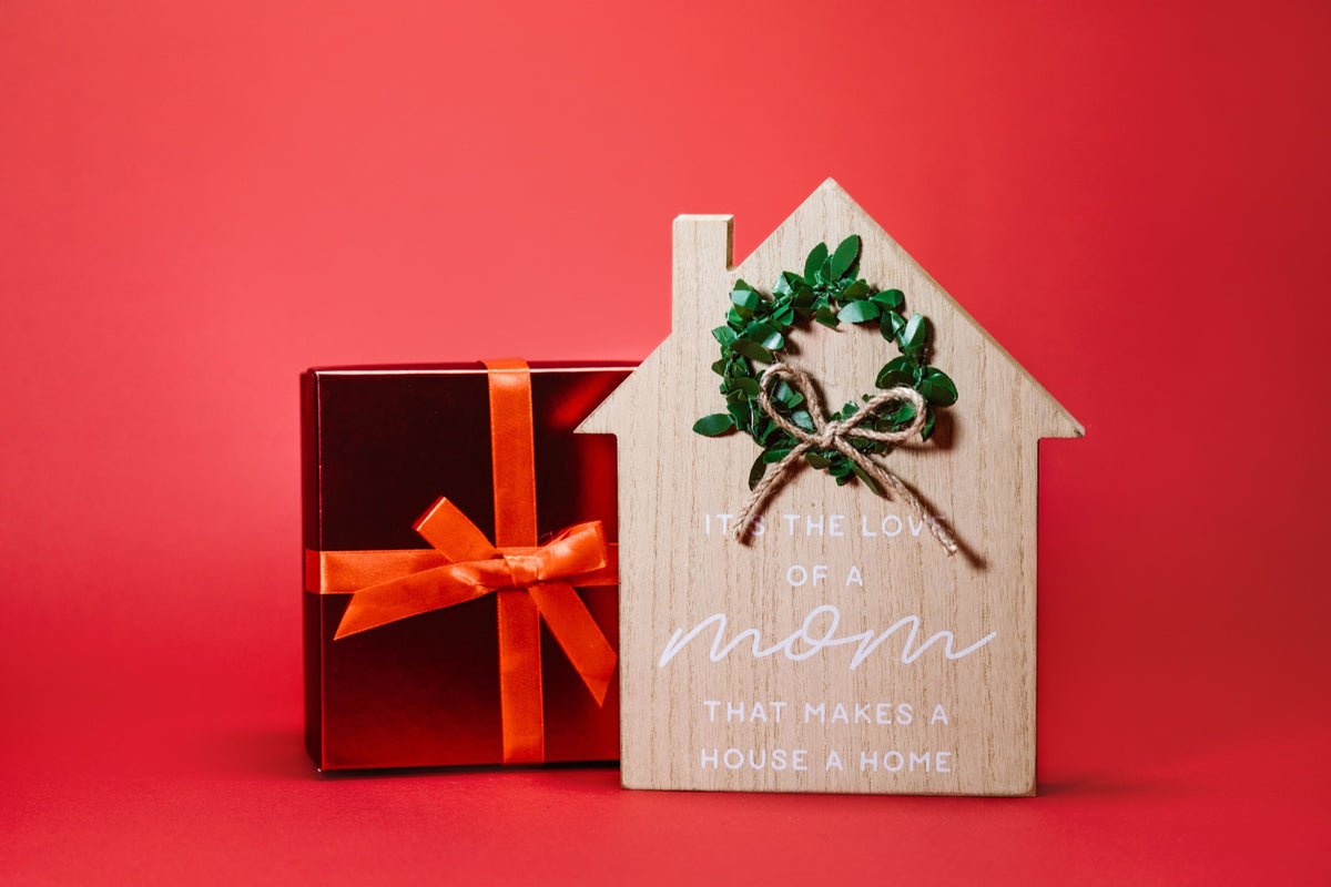 A wrapped gift box beside a small wooden house sign reading a message about the love of a mom